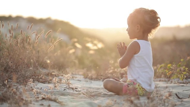 Little cute girl in white shirt meditates at sunset sitting on the sand