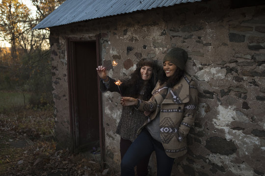 Two female friends leaning against outbuilding playing with sparklers