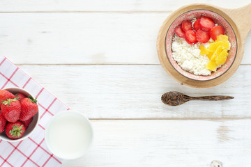 Cottage cheese with berries and dried mango, strawberry, cup of milk, wooden spoon and linen napkin on a white wooden table. Healthy breakfast. Top view.
