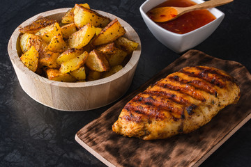 Grilled chicken breast on wooden board and grilled potato served with vegetables on black background