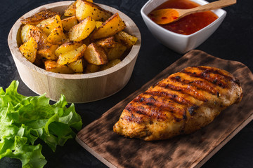 Grilled chicken breast on wooden board and grilled potato served with vegetables on black background