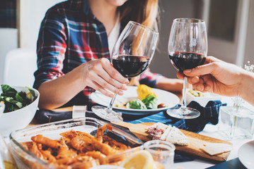Happy couple cheering with glasses of red wine, romantic atmosphere, hipsters making cheers at home, healthy food on the table, celebrating and enjoyment concept