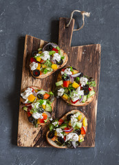 Greek style bruschetta on a wooden cutting board, on dark background, top view. Delicious appetizers for wine or a snack