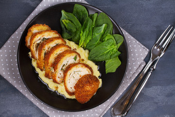 Chicken Kiev on mashed potato with spinach. Breaded chicken breast stuffed with herbs and butter on black plate. View from above, top studio shot