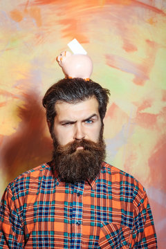 Frown bearded man holding pink ceramic piggy bank on head