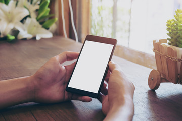 Mockup image of hands holding black mobile phone with blank white screen on vintage wooden table in cafe