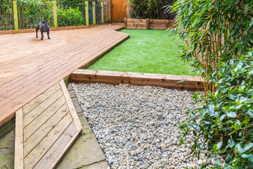 A section of a residntial garden, yard with wooden decking, patio over a fish pond, a section of artificial grass and an area of stone pebble. There is a bamboo plant and a dog in the garden. - obrazy, fototapety, plakaty