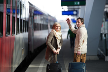 Senior couple on train station pulling trolley luggage, waving.