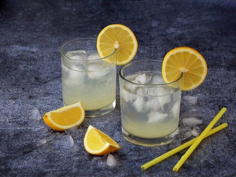 Two glasses of cold homemade lemonade with lemon slices, ice cubes and straws on dark background. Copy space