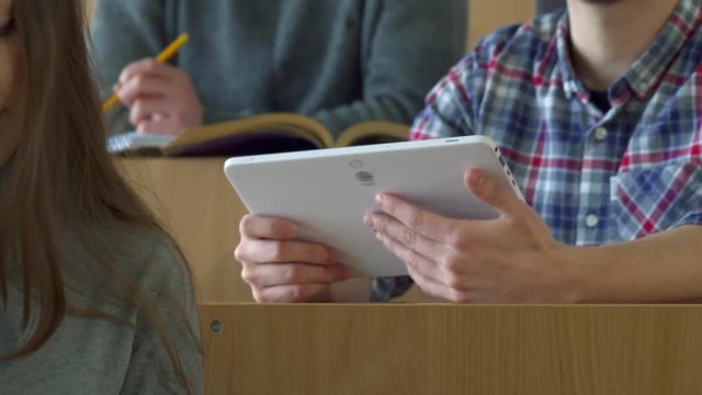 Young Male Student Showing His Classmate Something On His Tablet At The College. Caucasian Bearded Guy Turning To His Friend At The Lecture Hall. Camera Tilting From Front Rows With Undergraduate To