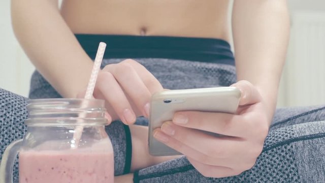 Young woman is drinking a smoothies and using a fitness app on her smartphone after a workout.