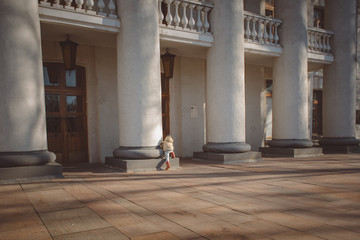 Obraz premium child in a beige jacket, rubber boots and hat in theater columns in the early spring on a sunny day walks