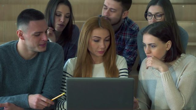 Group of students looking at laptop screen at the college. Young caucasian undergraduates discussing something on computer. Young people sitting at the lecture hall