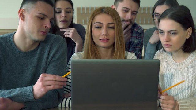 Group of caucasian students disscussing something on laptop at the college. Brunette bearded guy sitting on the rare row between two attractive female classmates. Pretty redhead girl sitting at the