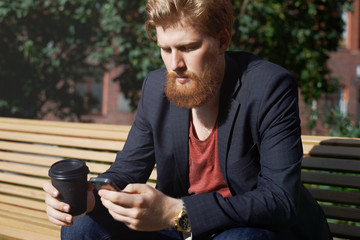 Sad beard man use phone to find new job. Modern hipster drink coffee and surf internet outdoor after study. Concentrated look while playing internet game online on sunny day outdoor.