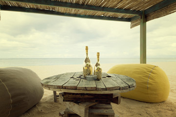 Two bottles of champagne on an old wooden table with pouf seats for romantic dinner for honeymoon copules on a tropical beach