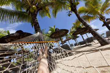 Female legs on hammock on tropical beach with palm leaf thatch roofing umbrellas and palm trees in the background