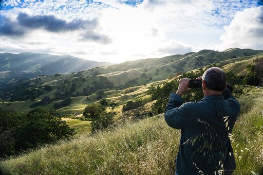 Man standing on grassy hill takes photo of sunset in tree covered valley