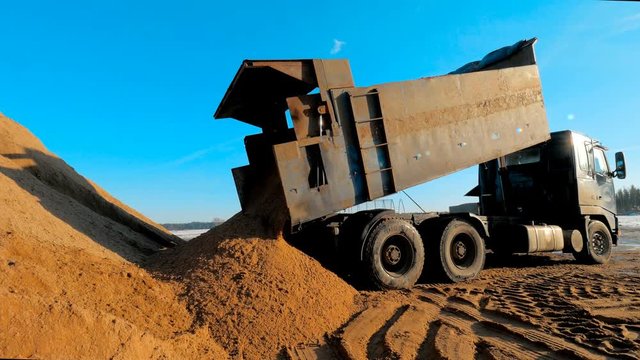 Dump truck unload soil at construction site. Big three-axle tipper truck pours out the sand at quarry career or open pit.