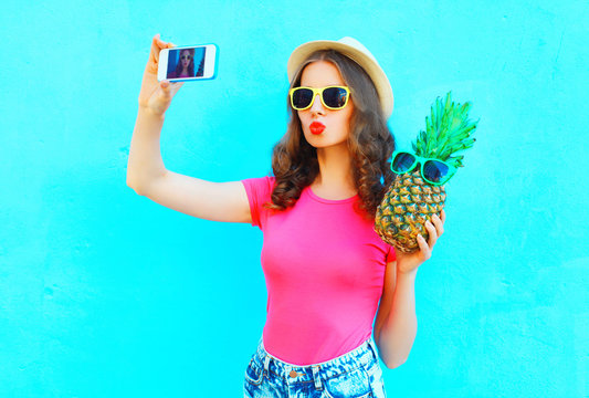 Fashion pretty woman taking picture self portrait on smartphone with pineapple wearing straw hat over colorful blue background