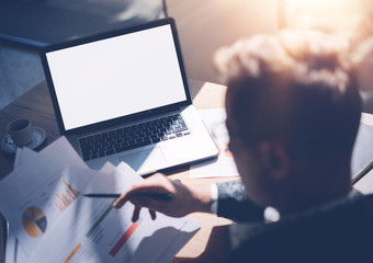 Adult banking analyst in eyeglasses working at sunny office on laptop while sitting at wooden table.Businessman analyze document in his hands.Graphs and diagramm on notebook screen.Horizontal mockup.