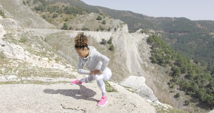 Young female wearing sportswear and stretching body while standing on rock cliff with mountains on background.