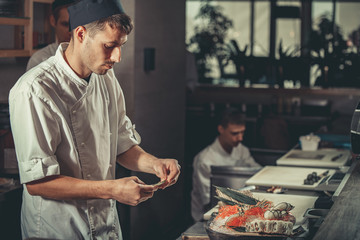 Two young white chefs dressed in white uniform decorate ready dish in restaurant. They are working on maki rolls. Preparing traditional japanese sushi set in interior of modern professional kitchen