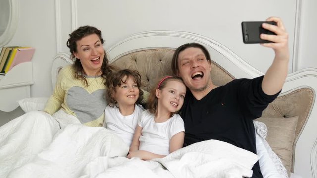 Positive Family Selfie with Mother, Father and Two Children Lying on the Bed and Wearing Pajamas. Handsome Man is Taking a Photo by His Smartphone.