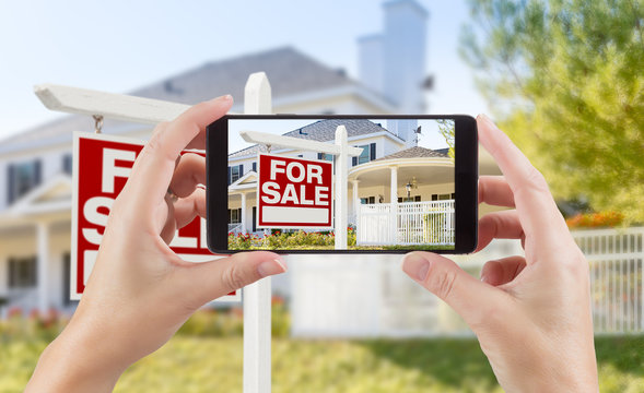 Female Hands Holding Smart Phone Displaying Photo of For Sale Real Estate Sign and House Behind.