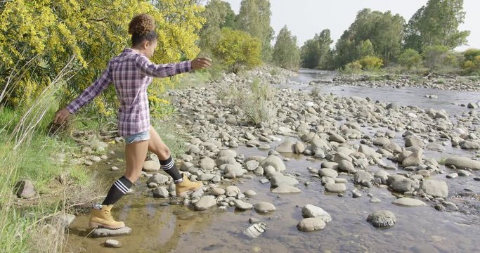 Young fit woman in casual outfit walking on rocks through small creek with green tress on background. 