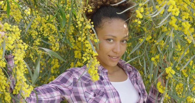 Smiling beautiful woman in casual outfit standing among trees brunches with yellow flowers and smiling at camera. 