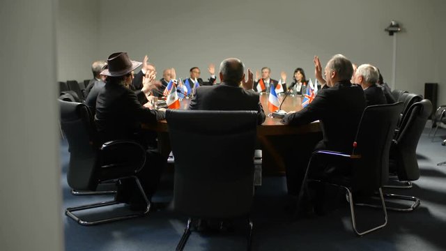 Delegates from different countries sit at the round table and vote unanimously. Policians raise up hands and approve the new law. The result of successful negotiations at international summit.