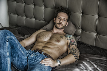 Shirtless sexy male model lying alone on his bed in his bedroom, looking at camera with a seductive...