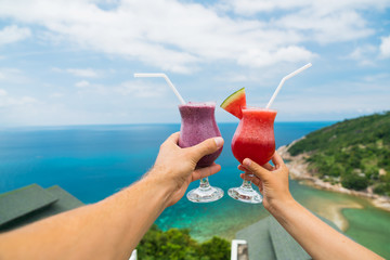 Fruit shake from fresh watermelon and blueberries on the outstretched hand of a man and a woman against a background of a tropical landscape. Luxurious vacation.