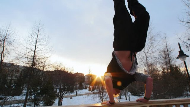 Work-out athlete - blonde man stands on hands in winter park at sunset