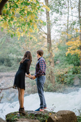 Fototapeta premium pair of beautiful brunette with a guy holding hands in nature at the waterfall fall trees