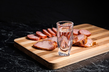 Shot glasses with vodka, slices of smoked meat and smoked sausage on wooden cutting board.