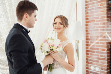 Groom gives wedding bouquet to the bride standing before panoramic window