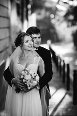 Tall groom hugs elegant bride from behind while they stand on summer street