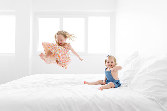 Two smiling sisters playing and jumping on a white bed