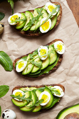 Avocado sandwich on dark rye bread made with fresh sliced avocados with spinach, guacamole, arugula and quail eggs on parchment paper. View from above. Healthy breakfast concept.