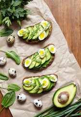 Spring food. A whole grain bread toast sandwiches with avocado, spinach, guacamole, arugula and quail eggs on parchment and dark wooden table. Healthy breakfast or lunch concept. Flat lay