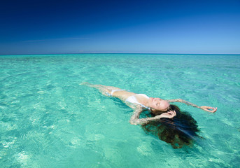 Beautiful young brunette woman with long hair dressed in white swimsuit lies on the crystal azure water of caribbean sea