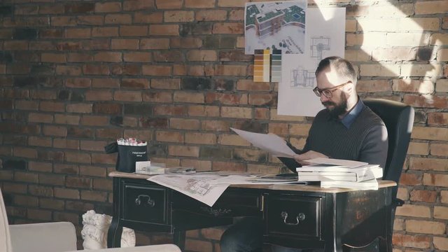 Man is working at the vintage table. Architect sitting at desk and working, there is blueprints and model house on his table. A stylish architect works in an office with a brick wall lit by sunlight