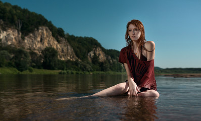 Yung beautiful redhead girl in the burgundy shirt sitting in the river