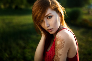 Naklejka premium Portrait of a red-haired girl with a mehendi on her shoulder against a background of greenery in the sunset light