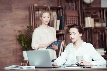 Young businesswoman with digital tablet looking at colleague using laptop