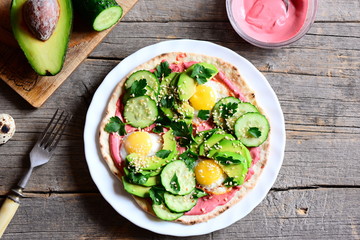 Home tortilla piled high with fried quail eggs, avocado, cucumber, sesame seeds and parsley and lathered with beetroot hummus. Half avocado, beetroot hummus in bowl, fork on a wooden table. Top view 