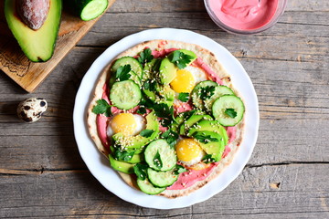 Homemade tortilla piled high with fried quail eggs, avocado, cucumber, sesame seeds and parsley and lathered with beetroot hummus. Avocado half, beetroot hummus in bowl, quail egg on a wooden table