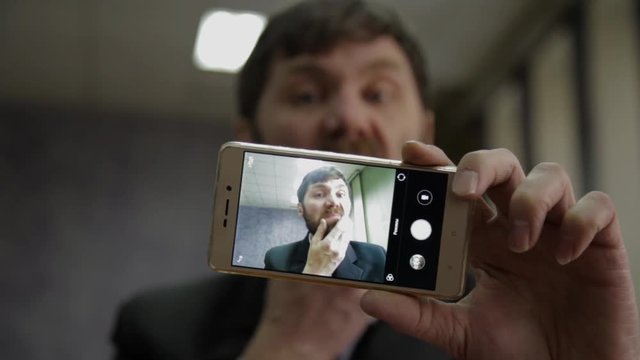 Busy office worker cranks and looking into phone camera. funy businessman picks finger in nose, strokes his beard